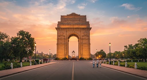 A detailed, vibrant street-level view of India Gate in New Delhi during golden hour. The majestic archway stands tall against a warm, saffron-tinted sky. The architectural details are crisp, and the surrounding space is uncluttered, emphasizing the monument's grandeur. The lighting creates a sense of warmth and invitation, perfect for an editorial travel aesthetic.